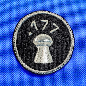 May include: A circular black patch featuring a silver keyhole design and the number .177 above it. The patch has a silver border, set against a blue textured background.