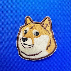 Può includere: Toppa ricamata raffigurante il meme Doge, un cane Shiba Inu con un'espressione sorridente. La toppa ha un contorno marrone, con un viso marrone e bianco, su sfondo blu.