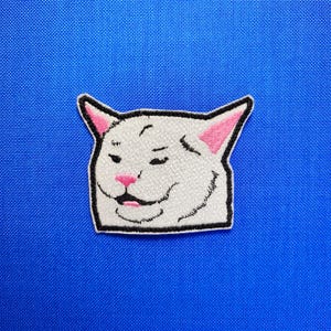 Op de afbeelding: Geborduurde patch met de meme van een witte kat met een sceptische uitdrukking. De kat heeft roze binnenoren, een roze neus en een zwarte omtrek. De patch staat op een blauwe achtergrond.