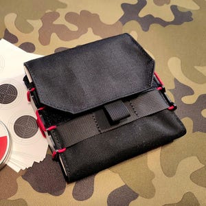 May include: Black tactical pouch with red stitching and a flap closure. The pouch has a black strap with a buckle. Target practice sheets are visible next to the pouch. The background is a camouflage pattern.