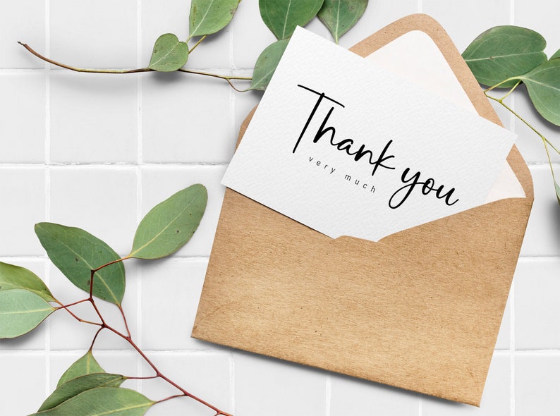 May include: A white card with the text "Thank you very much" in black calligraphy, inside a brown paper envelope. The card and envelope are on a white tiled surface with green leaves.