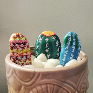 May include: Three painted rocks in a brown ceramic pot, each depicting a different cactus with white, pink, green, blue, yellow, and red colors.
