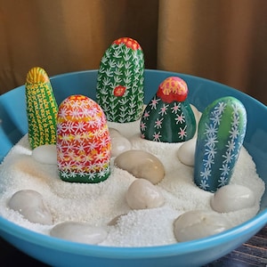 May include: A blue bowl filled with white sand and painted rocks shaped like cacti. The cacti are painted in various colors, including green, yellow, red, and white.