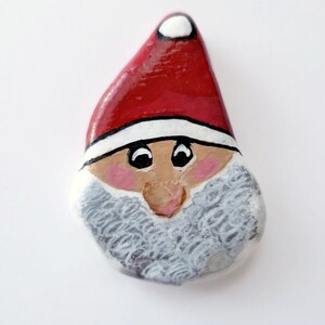 May include: Hand-painted rock art of Santa Claus with a red hat, white beard, and brown eyes.