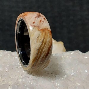 Pode incluir: Um anel de madeira com interior preto. O anel apresenta um padrão de grão de madeira natural com tons de bege, castanho e creme. O anel é exibido em uma superfície cristalina branca.
