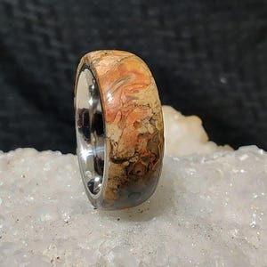 Pode incluir: Um anel de madeira com um padrão único e colorido, inserido em uma banda de aço inoxidável. O anel é uma peça de destaque com um visual natural e orgânico.