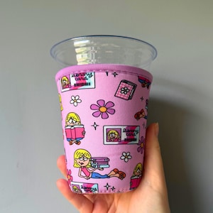 May include: A pink neoprene cup sleeve featuring a repeating pattern of cartoon characters reading books, library cards with the text library card, and white and purple flowers. The sleeve is designed to fit around a clear plastic cup.
