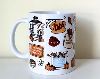 May include: A white ceramic mug featuring a colorful graphic print of autumn-themed illustrations, including a gazebo, a Luke's sign, a Stars Hollow Books tote bag, pumpkins, a cinnamon roll, and an orange ticket labeled Stars Hollow Autumn Festival.
