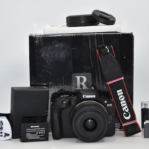 Op de afbeelding: Een zwarte Canon EOS digitale camera met lens, een zwarte camerariem met rode accenten, een oplader, een accu, een witte stroomadapter, een zwarte doos, een geheugenkaart en een kaartlezer. Het Canon-logo staat op de camerabehuizing.