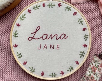 May include: A circular wooden embroidery hoop containing off-white fabric with the name Lana Jane stitched in dark red thread. The text is surrounded by a circular border of small embroidered green leaves and tiny red flower buds.