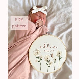 May include: An embroidery hoop featuring the name "ellie amelia" in brown thread, surrounded by embroidered daisies and a butterfly. The words "PDF pattern" are in a box in the upper left corner. The hoop is held near a sleeping baby.