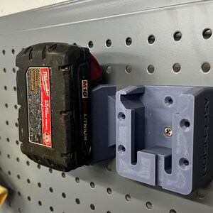 May include: A gray 3D-printed battery holder for a Milwaukee M18 battery pack, mounted on a gray pegboard. The holder is labeled "ASCENSION". The battery pack is black with red accents.