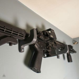 Può includere: Un fucile nero con un mirino a punto rosso montato sulla parte superiore è fissato a una parete con due ganci di plastica grigi. Il fucile ha un calcio nero e un'impugnatura a pistola nera.