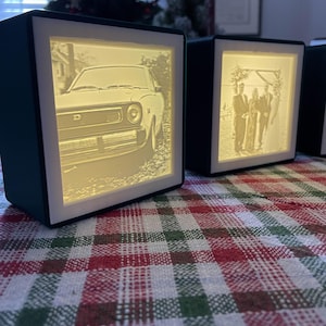 May include: Two dark teal illuminated photo boxes with white frames. One box displays a classic car, the other a wedding photo. The boxes sit on a red, green, and white plaid tablecloth. The boxes are lit from within.