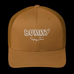 May include: A brown trucker cap with a mesh back and a curved peak. The front of the cap displays the word "BUMMY" in white, stylized lettering above the words "Spicy Jame" in a cursive font.