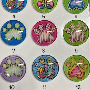 May include: A collection of twelve round, colorful paw print designs. Each paw print features a unique pattern and color scheme, including blue, green, pink, and yellow. The designs are likely for crafts or decorative purposes.
