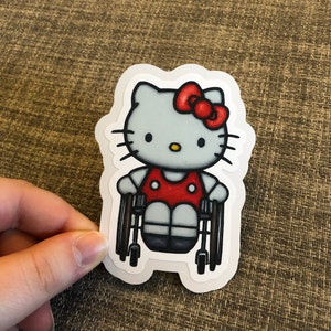 Könnte beinhalten: Ein weißer Hello Kitty-Charakter mit einer roten Schleife sitzt in einem Rollstuhl. Der Rollstuhl ist schwarz mit roten Rädern. Der Charakter trägt ein rotes Kleid mit weißen Punkten.