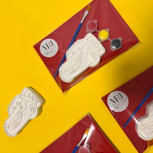 May include: Several paint-your-own car kits on a yellow background. Each kit includes a white car-shaped plaster figure, paint pots in red, yellow, and black, and a paintbrush. The kits are packaged in red cellophane.