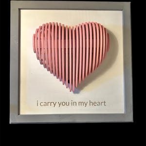 May include: A pink, layered heart sculpture within a gray framed artwork. The heart is made of vertical wooden slats. The artwork includes the phrase "i carry you in my heart" in a light gray font.