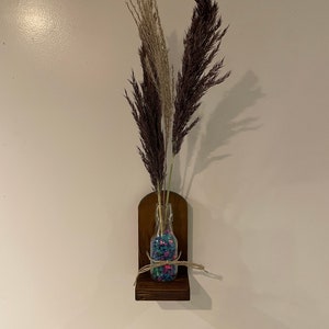 May include: A wooden wall shelf with a small glass vase filled with colorful pebbles and dried pampas grass.