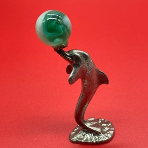 May include: A silver dolphin figurine holding a green and white glass marble. The dolphin is standing on a textured base.