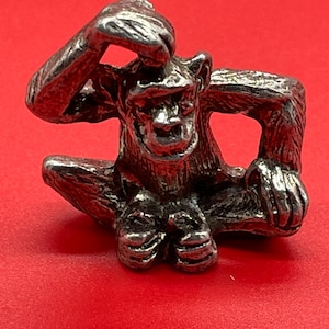 May include: A silver figurine of a monkey sitting with its hand on its head. The monkey is detailed with fur and facial features.
