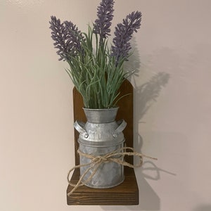 May include: A small wooden shelf with a metal milk can vase filled with purple lavender flowers. The shelf is mounted on a white wall.