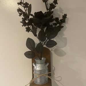 May include: A decorative wall-mounted floral arrangement. It features a galvanized metal milk jug vase with a jute twine bow, set on a dark brown wooden shelf. The vase holds black artificial flowers and foliage, creating a rustic aesthetic.