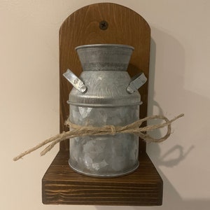 May include: A rustic wooden shelf with a galvanized metal milk can, tied with twine, mounted on the wall.