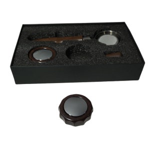 Puede incluir: Una caja negra con un asa de madera marrón, un filtro de metal plateado y una pequeña perilla de madera marrón. La caja está forrada con espuma negra.