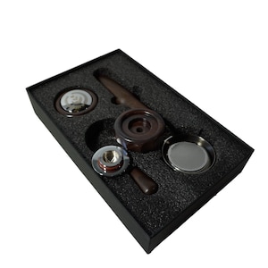 Puede incluir: Una caja negra con un interior de espuma que contiene un juego de herramientas de espresso. Las herramientas incluyen un tamper, un portafiltro, una herramienta de distribución y un filtro ciego. Las herramientas están hechas de metal y madera.