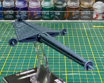 May include: A gray plastic spaceship model with a long, thin body and a large, flat wing. The model is mounted on a clear plastic stand and is sitting on a green grid.