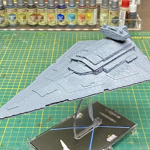 May include: A grey model Star Destroyer spaceship, with detailed surface features, mounted on a clear stand. The model is set against a green cutting mat, with paint bottles in the background.