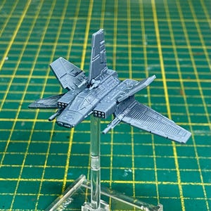 May include: A gray miniature spaceship model with detailed wings, engines, and a cockpit. The model is mounted on a clear plastic stand, set against a green grid background. The spaceship has multiple missile launchers and intricate paneling.