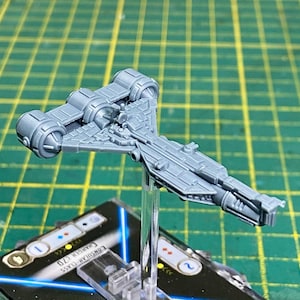 May include: A gray plastic model of a spaceship, featuring detailed paneling and cylindrical engines. The model is mounted on a clear stand, with a game card visible below. The spaceship is a Consular-Class Charger C70.