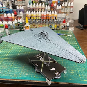 May include: A gray plastic model of a Star Destroyer spaceship featuring a triangular hull with intricate surface paneling and a central command tower, mounted on a clear plastic stand attached to a black rectangular base with printed graphics.