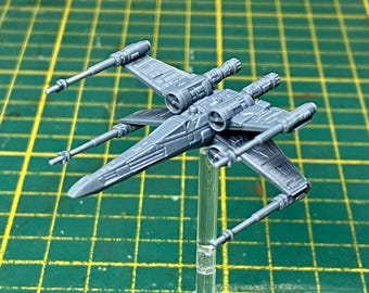 Puede incluir: Maqueta de plástico gris de un caza estelar X-wing, una nave espacial ficticia de una franquicia de ciencia ficción. El modelo está montado sobre un soporte transparente, con las alas en posición abierta. El modelo es detallado.