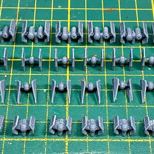 Puede incluir: Una colección de pequeñas maquetas de plástico gris de naves espaciales dispuestas sobre una superficie de cuadrícula verde. Los modelos incluyen varios tipos de cazas estelares y naves capitales.