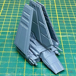 Puede incluir: Modelo gris de una nave espacial con alas anguladas y un cuerpo central. Las alas están abiertas, revelando detalles interiores. El modelo está sobre una superficie de cuadrícula verde.