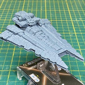Puede incluir: Modelo en miniatura gris y detallado de una nave espacial, posiblemente para un juego de mesa. La nave tiene un diseño complejo con múltiples niveles y detalles intrincados en la superficie. Montado sobre un soporte transparente.