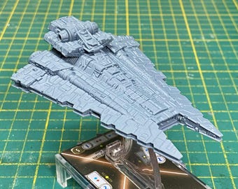 May include: A gray, detailed miniature starship model, possibly for a tabletop game. The ship has a complex design with multiple levels and intricate surface details. It is mounted on a clear stand above a game card.