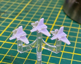 Op de afbeelding: Drie kleine, lichtpaarse, plastic model ruimteschepen worden getoond op een transparante plastic standaard. De standaard heeft drie armen die uitsteken en de ruimteschepen op hun plaats houden. De standaard staat op een groen rasteroppervlak.