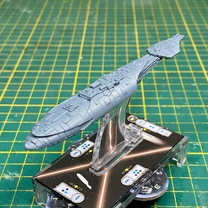 May include: A gray spaceship model with intricate detailing, mounted on a clear stand. The model is set against a green grid background, showcasing its design and structure. The base features game-related symbols.