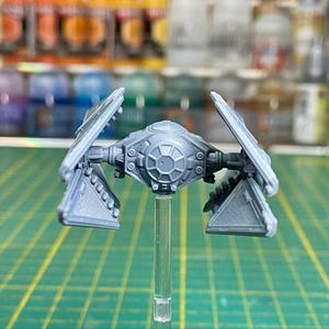 Op de afbeelding: Een grijs TIE-jagermodel met een gedetailleerd ontwerp. Het model staat op een transparante plastic standaard.