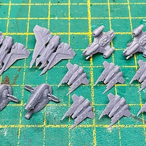Könnte beinhalten: Eine Sammlung grauer Miniatur-Raumschiffmodelle, darunter verschiedene Designs von Kampfflugzeugen und größeren Raumschiffen. Die Modelle sind auf einer grünen Rasteroberfläche angeordnet und zeigen ihre komplizierten Details und ihre futuristische Ästhetik.