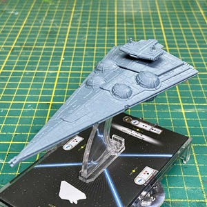 May include: A gray model of a starship, featuring a detailed design with multiple turrets and a bridge. The model is mounted on a clear stand above a black game card with white and blue accents. The ship is a light gray color.