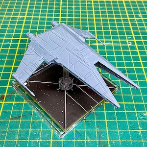 May include: A gray plastic model of a spaceship with a detailed design. The spaceship is on a black base with white lines.