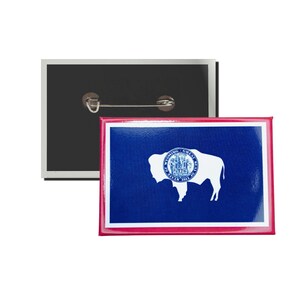 May include: A Wyoming state flag pin with a white buffalo silhouette and the state seal in the center. The pin is on a black background with a silver pin back.