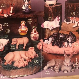 May include: A collection of hand-painted ceramic figurines depicting farm animals, including cows, pigs, cats, dogs, and a goose. The figurines are displayed on a wooden shelf with a variety of backgrounds, including a hay bale, a pillow, and a basket. The figurines are all in a relaxed pose, suggesting a theme of "lazy days".