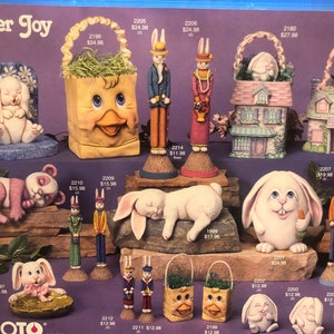 Può includere: Una collezione di statuine di coniglietti di Pasqua in ceramica, tra cui un coniglietto che dorme, un coniglietto che tiene una carota e un coniglietto con un cestino. Ci sono anche altre statuine a tema pasquale, come una borsa a forma di anatra, una casa e un orso panda.